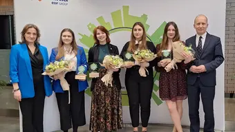 Image of six people standing in front of a photo wall, 4 female students with flower bouquets and two professors
