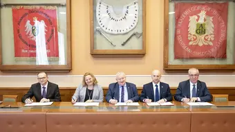Na zdjęciu moment podpisania umowy. Sygnatariusze siedzą za stołem prezydialnym, trzymając w dłoniach długopisy. Przed nimi dokumenty. Zdjęcie wykonane w sali konferencyjnej AGH.