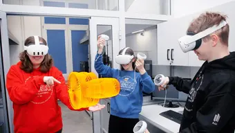 Zdjęcie wykonane w laboratorium. Troje młodych ludzi ma założony na głowy specjalistyczny sprzęt przypominający zabudowane okulary VR. W dłoniach trzymają rodzaj bezprzewodowych uchwytów, którymi manipulują. Pomiędzy stojącymi w laboratorium młodymi ludźmi widoczny jest wygenerowany przez nich trójwymiarowy obraz przedmiotu w kolorze żółtym.