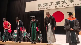 Zdjęcie z uroczystości rozdania dyplomów. Na scenie stoją osoby, które odebrały dyplom. Są ubrane w ceremonialne stroje japońskie. Po prawej stronie stoi prof. Monika Motak w japońskiej todze dziekańskiej.