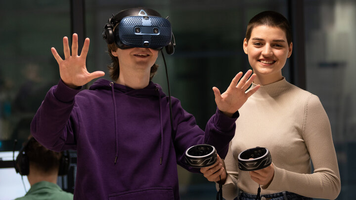 Zdjęcie przedstawia dwoje młodych ludzi. Mężczyzna po lewej ma na twarzy okulary VR i wyciągnięte przed siebie dłonie. Kobieta po jego prawej przekazuje mu dwa manipulatory.