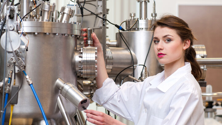 Zdjęcie przedstawia kobietę ubraną w biały fartuch w laboratorium. Po jej lewej stronie cylindryczna aparatura wykonana ze szczotkowanego metalu. W górnej części aparatury widać ogromną ilość doprowadzonych do niej przewodów. 