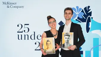 The photo shows a woman in a long black dress and a man in a black suit and white shirt, they are both smiling. In the background is a wall with the inscription “25 under 25”.