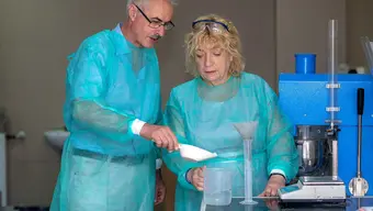 Zdjęcie przedstawia dwóch pracowników naukowych w niebieskiej odzieży ochronnej podczas pracy w laboratorium. Mężczyzna w okularach po lewej stronie wsypuje łopatką odczynnik chemiczny do zlewki wypełnionej w połowie wodą, którą trzyma kobieta po prawej stronie. Na stole laboratoryjnym znajduje się też menzurka z włożonym lejkiem oraz elektroniczna waga. Po prawej stronie stoi niebieski sprzęt laboratoryjny.