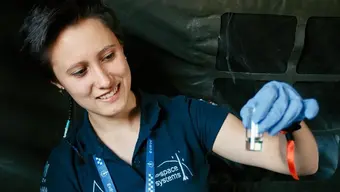 The photo shows a young woman in an AGH Space Systems T-shirt who is examining a transparent fluid in a glass container that she is holding up to her face with her left arm. She is also wearing blue disposable gloves and a lanyard.