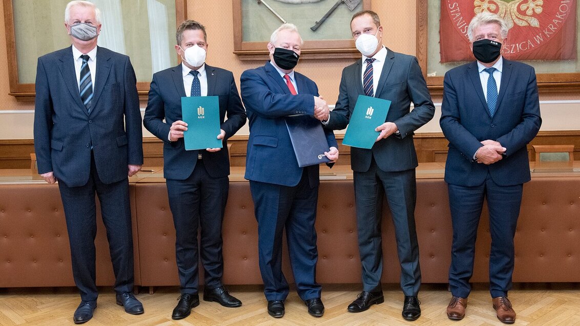 Zdjęcie zrobione w sali konferencyjnej, przed stołem prezydialnym. Przedstawia pięciu mężczyzn stojących obok siebie w rzędzie. Mężczyźni są elegancko ubrani w ciemne garnitury, białe koszule i krawaty. Usta i nos mają zakryte maseczkami. Wzrok kierują do aparatu, w ich oczach widać zadowolenie. Trzech stojących pośrodku trzyma w dłoniach eleganckie teczki z logo AGH z podpisanym porozumieniem, dwaj z nich podają sobie dłonie w geście gratulacji. Za mężczyznami, na ścianie znajdują się trzy gabloty w których zamieszczone są godło AGH oraz insygnia władzy: berło i topór ceremonialny.