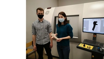 Na zdjęciu stoją obok siebie: Grzegorz Kaczmarczyk (po lewej) i doktor Justyna Orłowska (po prawej). Ich usta i nosy zakrywają maseczki. Doktor Orłowska trzyma w dłoniach fragment poroża. Za plecami stojących osób widać boczną ścianę tomografu w kolorze jasnobeżowym. Po prawej stronie stojących osób znajduje się ekran komputera, klawiatura oraz myszka komputerowa i panel sterujący tomografem z przyciskami. Na ekranie komputera widać tomograficzne zdjęcie poroża.