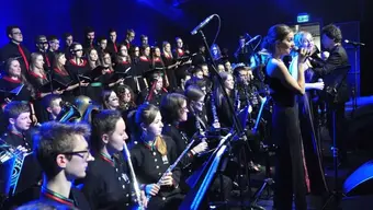 Zdjęcie przedstawia wnętrze z przyćmionym, niebieskim światłem. Po prawej stronie - dwie solistki ubrane w długie, czarne suknie. Każda z nich trzyma w ręku mikrofon. Za nimi widać dyrygenta. Za solistkami i dyrygentem widoczne cztery rządy muzyków; pierwsze dwa - z miejscami siedzącymi, muzycy ubrani w czarne mundury (na stójkach przy szyi oraz na końcu rękawów - lamówki w kolorze czerwonym i zielonym) trzymają w dłoniach instrumenty dęte. W kolejnych dwóch rzędach stoją chórzystki i chórzyści ubrani w czarne stroje z czerwonymi elementami.