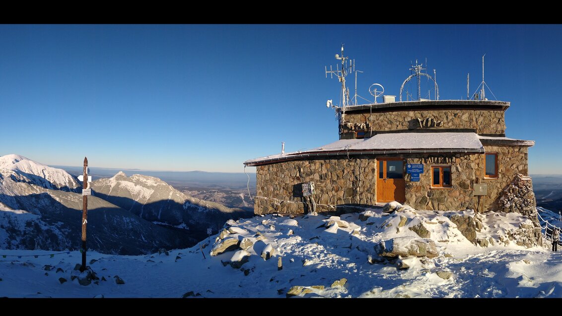 Zdjęcie przedstawia budynek Wysokogórskiego Obserwatorium Meteorologicznego IMiGW na Kasprowym Wierchu na tle panoramy Tatr. Budynek wykonany jest z szarego kamienia. Ziemia oraz dach budynku pokryte są cienką warstwą śniegu.