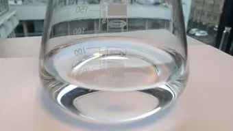 Szklana zlewka, która zawiera płynny, przezroczysty materiał powłokowy, używany podczas nanoszenia techniką dip-coatingu. Naczynie znajduje się na parapecie, za którym widoczne są budynki na terenie uczelnianego kampusu.