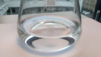 The photo shows a glass beaker that holds a liquid, transparent coating material applied using the dip-coating technique. The container is located on a window sill overlooking the AGH UST campus buildings.