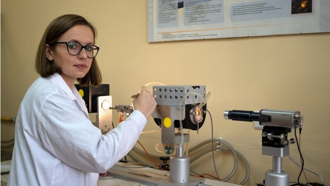 Na zdjęciu widoczna jest młoda kobieta w okularach i białym fartuchu, która stoi przed srebrnym mikroskopem wysokotemperaturowym