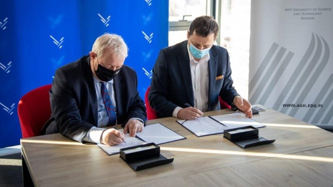 The photo shows two men in suits sitting at a table and signing the agreement. Behind them are two banners, one blue with STH logo and one white with the AGH UST logo.