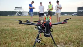 trawiasta powierzchnia na lotnisku; na pierwszym planie na trawie dron; na drugim planie, za dronem stoi obok siebie czterech młodych mężczyzn, piąty mężczyzna siedzi na trawie. Mężczyźni ubrani są w szorty i t-shirty. Jeden z nich, stojący po prawej stronie, ma na temblaku obandażowana prawą rękę. W tle – budynki lotniska, za nimi gęste pasmo drzew.