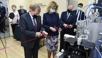 Zdjęcia przedstawia pomieszczenie. Po prawej stronie widoczny fragment aparatury badawczej. W środkowej części zdjęcia widoczne cztery stojące osoby, z których trzy (dwóch mężczyzn i kobieta) przecinają nożyczkami wstęgę rozwiniętą przed aparaturą. W tle, po lewej stronie widać cztery inne stojące osoby. Na ścianie widoczny napis: Wydział Odlewnictwa. 70 lat. Wszystkie osoby na zdjęciu mają maseczkami zakryte usta i nosy.