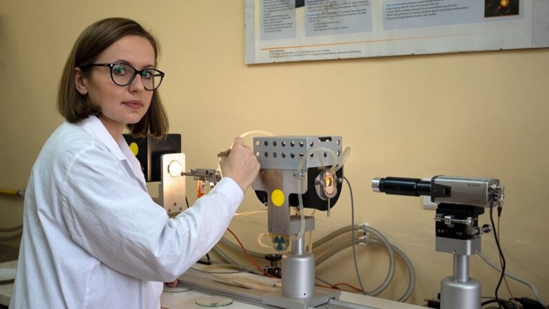 The photo shows a young woman in a lab coat standing in front of a silver high-temperature microscope.