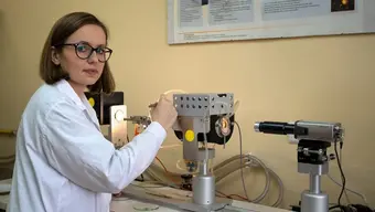 The photo shows a young woman in a lab coat standing in front of a silver high-temperature microscope.