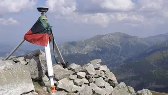 Po lewej znajduje się odbiornik GNSS. Odbiornik jest w kolorze stalowym. Stoi na trójnogu. Na urządzeniu umieszczona jest flaga AGH. Flaga składa się z poziomych pasów w kolorach zielonym, czarnym i czerwonym. Na drugim planie znajduje się grań górska. Widać granitowe skały pokryte miejscami zieloną roślinnością. Nad granią górską znajduje się błękitne niebo, które częściowo przykrywają chmury w kolorach białym i szarym.