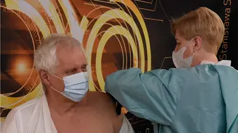 Pielęgniarka ubrana w fartuch medyczny szczepi w ramię starszego mężczyznę. Oboje mają twarze zasłonięte maseczkami medycznymi. Mężczyzna ma zdjętą z ramienia koszulę.