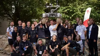 Grupa członków zespołu AGH Space Systems pozuje do wspólnego zdjęcia na zewnątrz. W centralnej części fotografii kilka osób unosi nad głowami łazik marsjański.