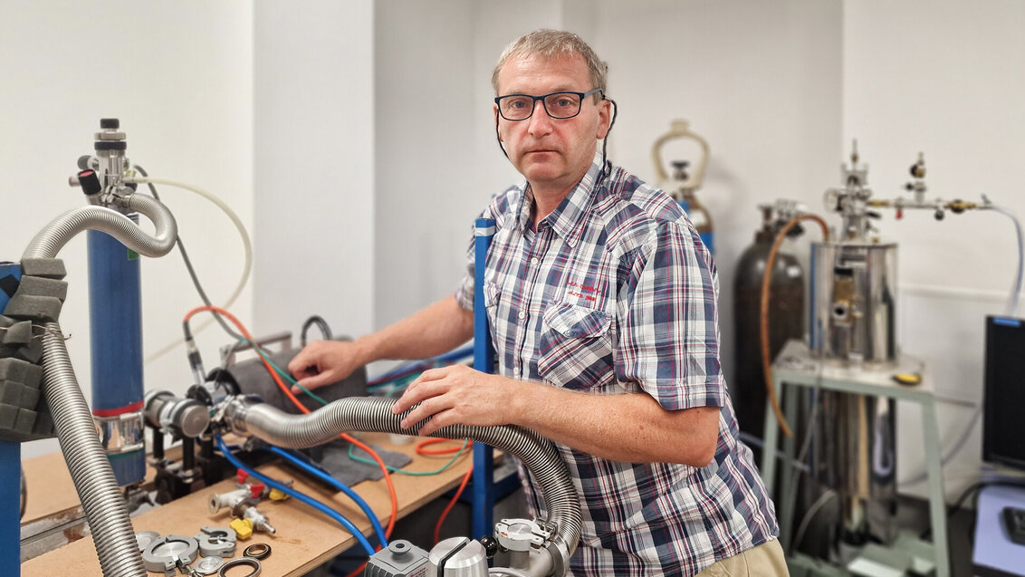 Mężczyzna w średnim wieku w okularach i kraciastej koszuli opiera się o rurę należącą do aparatury laboratoryjnej