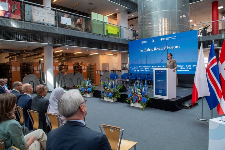 Speaker at a podium addressing an audience during the Baltic Rectors’ Forum, with national flags beside the stage and attendees seated in a large modern atrium.