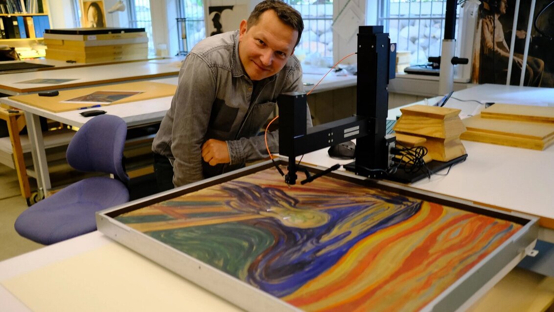 Tomasz Łojewski w pracowni. Naukowiec siedzi przy dużym blacie, na którym leży obraz "Krzyk: E. Muncha. Obok specjalistyczny sprzęt.