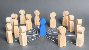 A colourful illustrative image of wooden figurines that mimic human figures. A blue figurine stands in the middle of a circle made of more than a dozen beige figurines. Each beige figurine is connected to the blue one with a white line running across the floor.