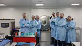 Grupa osób w niebieskich kombinezonach ochronnych stoi w laboratorium przed dużą komorą testową oznaczoną logo „ACS”. Na pierwszym planie widać stół z przyrządami i pojemnikami laboratoryjnymi.