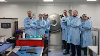 A group of seven young men in blue lab coats and hairnets standing with their arms crossed, smiling, in a laboratory setting. Behind them, there is a vacuum chamber with a small, round window.
