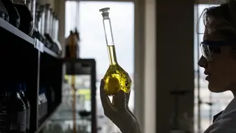 Na zdjęciu kobieta w laboratoryjnym ubraniu ochronnym (fartuch, okulary, rękawiczki) trzyma w dłoniach menzurkę wypełnioną cieczą.