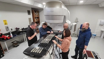 Zdjęcie wykonane w laboratorium. Grupa sześciu osób zebrana wokół specjalistycznego sprzętu wyglądającego jak długi, czarny panel z białymi przyciskami, do których podpięta jest plątanina kabli. Za panelem widać obudowę fantomu, która kształtem przypomina duży, pionowy sarkofag.