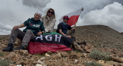 Trzy osoby siedzą na skalistym zboczu w górskim terenie, trzymają flagi Polski oraz AGH. W tle widoczne są chmury i rozległy, kamienisty krajobraz.