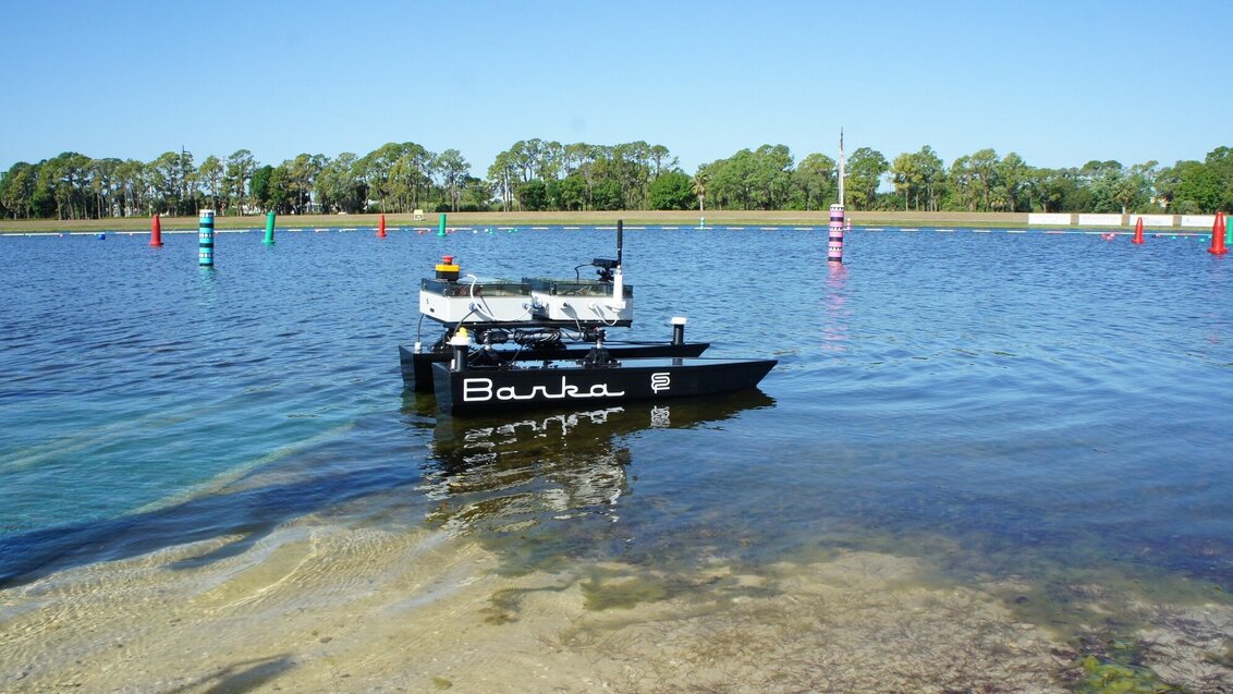 Na zdjęciu unosząca się na wodzie łódź autonomiczna "Barka". Ma dwa równoległe pływaki w kolorze czarnym połączone platformą w kolorze białym, na której umieszczone jest okablowanie i czujniki. Na jednym z pływaków widoczny napis "Barka".