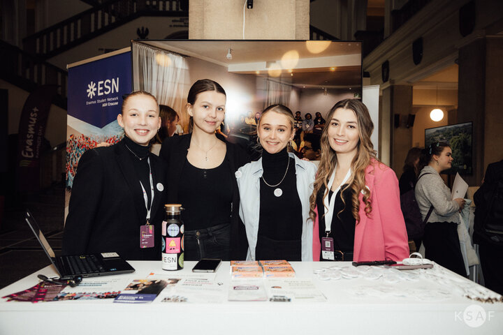 Zdjęcie czterech młodych kobiet stojących przy dużym stole. Prezentują Sieć Studencką Erasmus+. Na stole materiały marketingowe.