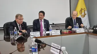 Three university representatives seated at a conference table during a formal meeting, with microphones, nameplates, and an institutional flag in the background.