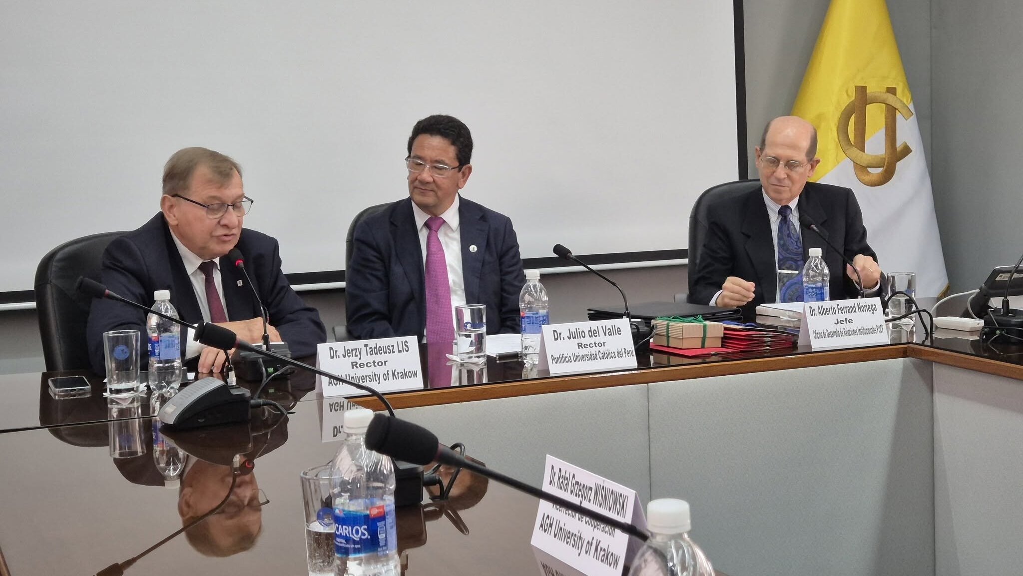 Three university representatives seated at a conference table during a formal meeting, with microphones, nameplates, and an institutional flag in the background.