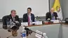Three university representatives seated at a conference table during a formal meeting, with microphones, nameplates, and an institutional flag in the background.