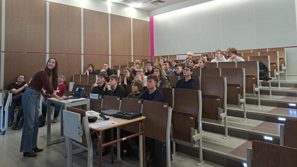 A group of students sitting in the audience in an auditorium. A woman st the front is displaying them something on a screen.