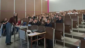 Sala wykładowa. Na pierwszym planie stoi kobieta przy laptopie, w tle uczniowie zasiadający w ławkach na sali wykładowej