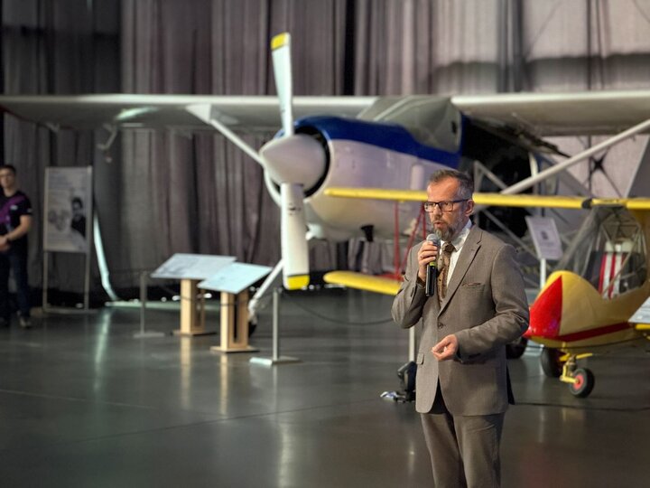 An elegant man is speaking into a microphone, with an aeroplane – part of the museum’s exhibition – in the background.