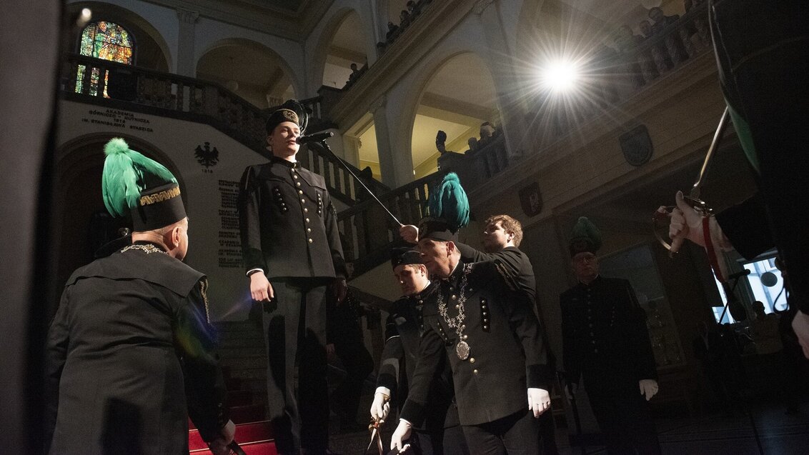 A ceremonial group in black mining uniforms with green-plumed hats inside a grand hall, with one person speaking into a microphone under dramatic lighting.