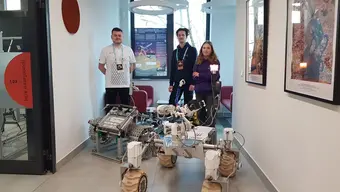 W korytarzu – łazik marsjański na sześciu kołach, wyposażony w moduły i czujniki. Obok łazika stoją trzy osoby.