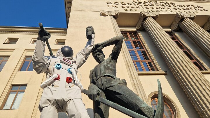 Na zdjęciu pomnik górników, za nim widoczny budynek główny AGH (A-0). Lwy górnik z pomnika ubrany jest w skafander astronauty, na głowie ma hełm.