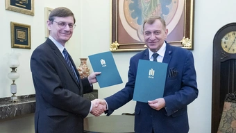 Two middle-aged men in suits shaking hands in a congratulatory gesture. The men are smiling and holding green folders with a gold AGH UST logo. In the background are antique furniture and paintings.