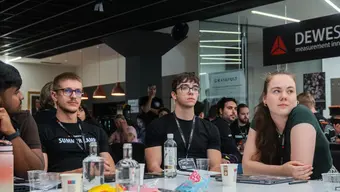 Uczestnicy szkolenia siedzą przy stole konferencyjnym i słuchają prezentacji. Na stole znajdują się m.in. laptopy oraz materiały szkoleniowe. W tle widoczne są stanowiska i oznaczenia firm technologicznych