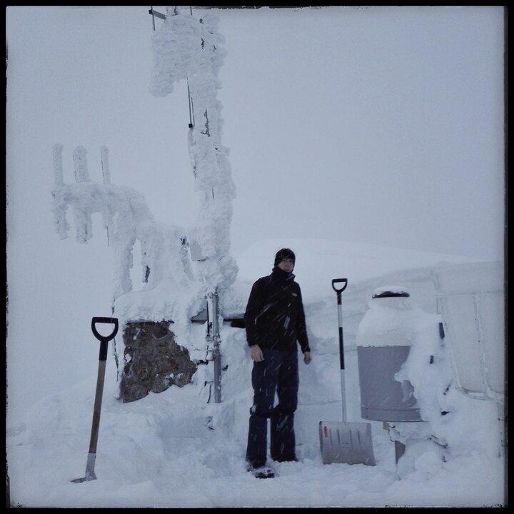 Mężczyzna stojący na tle Wysokogórskiego Obserwatorium IMiGW-PIB na Kasprowym Wierchu. Mężczyzna ubrany jest w czarną kurtkę i czarne spodnie. Budynek przysypany jest grubą warstwą śniegu.