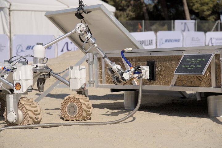 Photo of the rover Kalman performing a task, with the rover’s gripper manipulating a board in front of it.