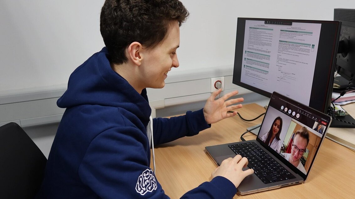Na zdjęciu student udzielający korepetycji. Siedzi przy biurku, przed nim ekran laptopa, na którym widać dwie twarze: uczennicy i ucznia. Na drugim monitorze widoczny skrypt korepetycji z zadaniami.
