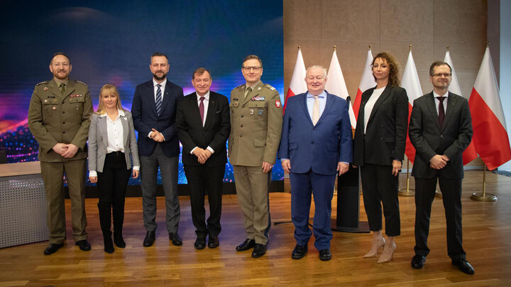 Mieszana, ośmioosobowa grupa pozuje do zdjęcia. Dwie kobiety w średnim wieku ubrane biznesowo. Wśród mężczyzn, dwóch nosi mundury wojskowe, pozostali w garniturach. W tle po prawej szpaler polskich flag narodowych.